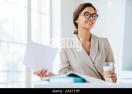 Fröhliche Geschäftsfrau liest Papierdokumente, konzentriert mit Lächeln Getränke Milchshake sitzt auf dem Desktop macht Geschäftsplan für Projekt. Weibliche Execut Stockfoto