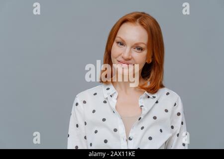 Gefiel Geschäftsfrau mit kurzen roten Haaren, Make-up, sieht positiv auf die Kamera aus, hat nach einem erfolgreichen Geschäftstreffen gute Laune, gekleidet in stilvollem Shi Stockfoto