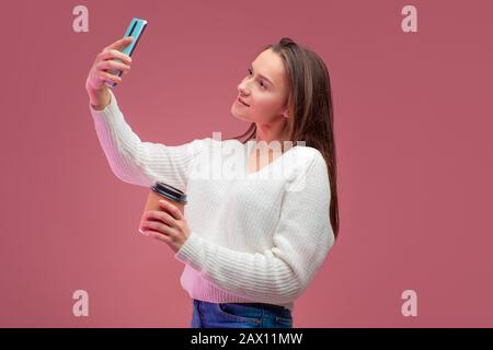 Süße junge Brunette in einem Pullover und Jeans hält einen heißen Kaffee und nimmt ein selfie auf ihr Smartphone. Stockfoto