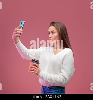 Süße junge Brunette in einem Pullover und Jeans hält einen heißen Kaffee und nimmt ein selfie auf ihr Smartphone. Stockfoto