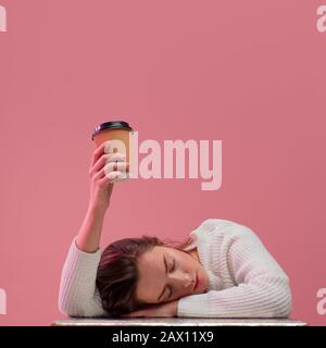 Ich brauche mehr Kaffee. Das schlafende Studenten-Mädchen schläft und zieht ihre Hand mit einer Tasse Kaffee nach oben und bittet um eine zweite Tasse Kaffee, pinkfarbenen Hintergrund. Stockfoto