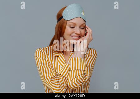 Süße Träume und gutes Ruhekonzept. Die hübsche Rotkopffrau dreht den Kopf beiseite, hält den Blick nach unten und berührt die Wange sanft, trägt gestreifte Pajama, Augenbinde, Stockfoto