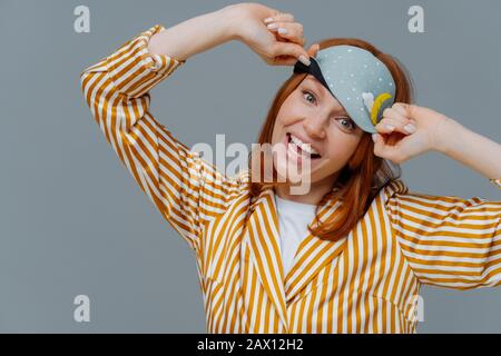 Die fröhliche Frau trägt einen blinden und gestreiften Schlafanzug, ist glücklich, erwacht in guter Laune, hat gesunde Schlafgewohnheiten, fühlt sich total entspannt an Stockfoto