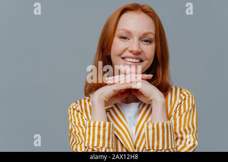 Nahaufnahme der lieblichen Frau hat foxy Haare, lächelt Zahnig an der Kamera, hält die Hände unter dem Kinn, ist im Homewear gekleidet, genießt eine ruhige häusliche Atmosphäre, Stockfoto