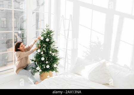 Positive junge Frauen posieren in der Nähe großer Fenster im gemütlichen Schlafzimmer, schmücken gern Weihnachtsbaum, tragen warme Kleidung, hat ein glückliches Lächeln im Gesicht, bereitet sich vor Stockfoto