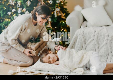 Schöne fürsorgliche Mutter bereitet sich auf kleine Kinder vor, die auf dem Boden in der Nähe des Weihnachtsbaums schlafen, überrascht den Urlaub, steht auf Knien nea Stockfoto