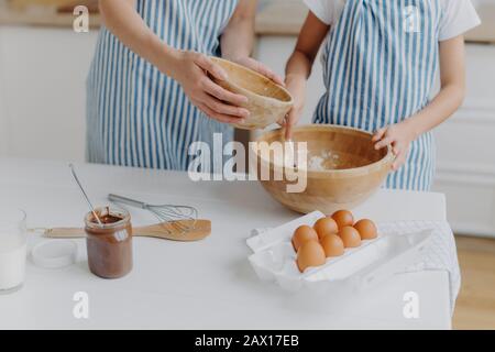 Zugeschnittenes Bild von Müttern und Töchtern Hände Mischen von Zutaten, um Teig zuzubereiten und leckeres Gebäck zu backen, stehen in der Nähe von Küchentisch mit Eiern, geschmolzenem Cho Stockfoto