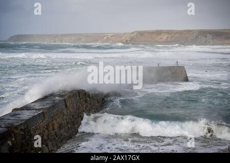 Wellen stürzen gegen die Hafenmauer in Sennen Cove nahe Land's End, Cornwall, wo immer noch eine gelbe Windwarnung in Kraft ist, während der Sturm Ciara über einige Teile des Vereinigten Königreichs zurückbleibt. Stockfoto