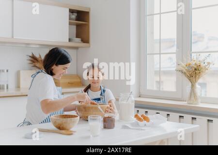 Die Frau mit Lächeln zeigt der kleinen Tochter, wie sie kochen kann, gibt kulinarische Lektion, kaufte verschiedene Produkte zum Kochen, trägt Schürzen und Lächeln Stockfoto