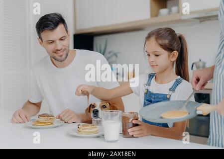 Foto des kleinen Mädchens in Denim Dungarees fügt Pfannkuchen Schokolade hinzu, frühstücken zusammen mit Papa und Hund, wie Mutter kocht. Familie in der Küche Stockfoto