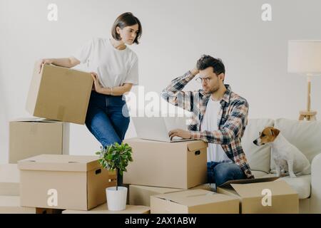 Unglücklicher männlicher Freiberufler in Brillen sitzt vor einem modernen Laptop, arbeitet ferngesteuert zu Hause, beschäftigt Frau steht in der Nähe mit Kartonbox, bewegt sich in neuem Apar Stockfoto