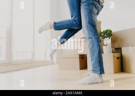 Glückliches Unbekanntes Paar bewegt sich in neuer Wohnung, Mann hebt Frau, trägt Jeans, Pose im leeren Raum mit Kartons herum, beginnt getrennt von Eltern r zu leben Stockfoto