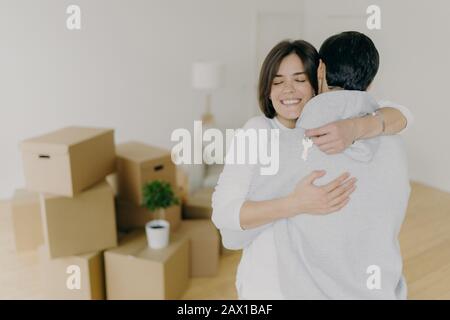 Fröhliche brünette lächelnde Frau umarmt ihren Mann mit Liebe, hält Schlüssel, kauft gemeinsam das erste Haus, kauft neues Grundstück, posiert in unmöbliertem Raumwit Stockfoto