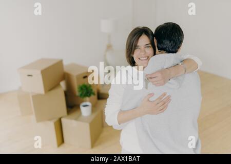 Innenschuss eines schönen Familienpaares umarmen sich mit Liebe, halten Schlüssel aus der neuen Wohnung, ziehen in der eigenen Wohnung, stellen sich in leerem hellen Wohnzimmer mit Fußboden Stockfoto