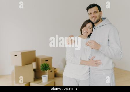 Liebespaar kauft sich ein eigenes Haus, umarmen und stehen sich eng aneinander, halten Schlüssel aus neuer Wohnung, Kartons im Hintergrund, haben sich begeistert f Stockfoto