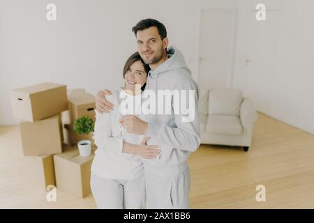 Jahrtausende Paare mit einem Lächeln, Umarmung und halten Schlüssel aus der eigenen Wohnung, kaufen ihr erstes Haus, bezogen im gemeinsamen Haus, stehen in leerem Raum mit Wagen Stockfoto