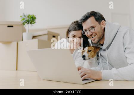 Überfreuliches Familienpaar lachen, während Sie Online-Shops auf einem modernen Laptop mit ihrem Hund ansehen, nach dem Auspacken von Kartons Pause machen, home renovatio Stockfoto