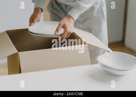 Zugeschnittenes Bild eines unbekannten gesichtslosen beschäftigten Mannes entpackt Kisten mit Geschirr, hält weiße Teller, bewegt sich in neuer Wohnung, tickt in der Küche auf, hat einen beweglichen Tag Stockfoto