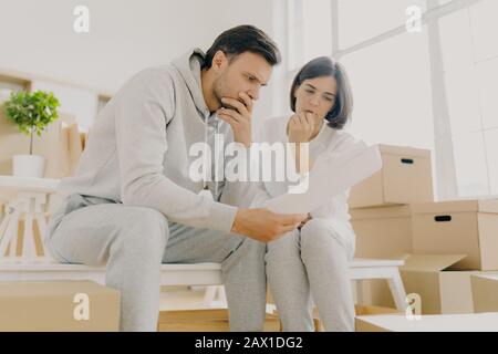 Besorgte Paare lasen Brief von der Bank, erhalten Benachrichtigung für die Zahlung von Rechnungen, haben trauriges Aussehen, sitzen auf der Bank in einem geräumigen Zimmer, gestresst, in Schwierigkeiten, hav Stockfoto