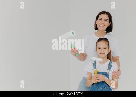 Lächelnde Mutter umarmt Tochtermaschine, steht mit Farbrollen, Farbwände im Haus, Mädchen hat schmutziges Gesicht, gekleidet in Denim-Overalls, Posen im Vernehmen Stockfoto