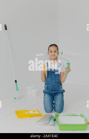 Glückliches kleines Mädchen in Denim-Overalls steht auf Knien, hält Farbrolle, umgeben mit Tabletts mit Farbe, Posen im weißen Raum, hat glücklichen Ausdruck, Stockfoto