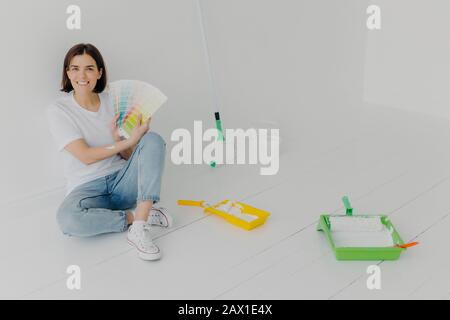 Glad Brunette Woman zeigt Farbmuster, gekleidet mit weißem T-Shirt und Jeans, wählt den Ton für die Wandsanierung, beschäftigt mit der Reparatur und mit dem Renovieren von und mit dem Stockfoto