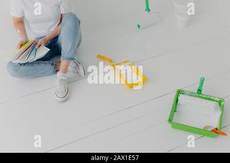 Zugeschnittenes Bild einer gesichtslosen Frau wählt Farbe aus Proben, wird Wände im eigenen Raum sanieren, trägt Turnschuhe, sitzt auf weißer Wand mit Tabletts und PA Stockfoto