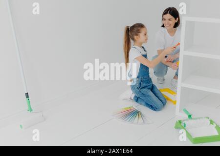 Geschäftige Mädchen und ihre lächelnde Mutter malen Schublade in weißer Farbe, verwenden Sie Pinsel oder Rolle, umgeben mit Farbproben, stellen Sie sich in weiß leeren Raum, denken Sie Stockfoto