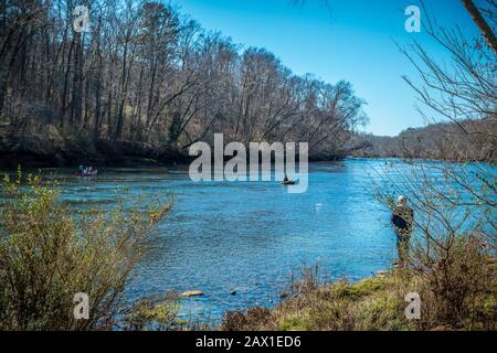 Ein Fischer, der im Wasser wate und ein Mann, der an der Küste angeln und mit einer Familie Kanufahren im Hintergrund jede Menge Aktivitäten auf dem Fluss austochen Stockfoto