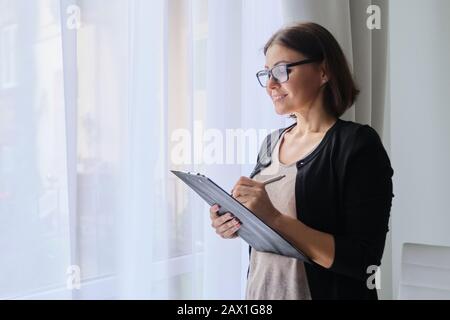 Reife Frau Lehrerin, die Notizen machte und in der Nähe von Fenster im Büro stand Stockfoto