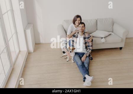 Portrait der fröhlichen brünette jungen Frau umarmt ihren Mann, der mit Stammhund auf dem Boden sitzt, in leerem, geräumigem Raum posieren, in neuem ap umziehen Stockfoto