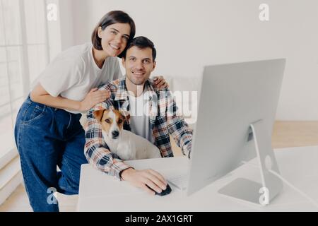 Foto des männlichen Freiberuflers arbeitet ferngesteuert am Computer mit Hund, seine liebevolle Frau umarmt und hilft bei einigen Fragen, stellt sich im großen Raum bei Workpl Stockfoto