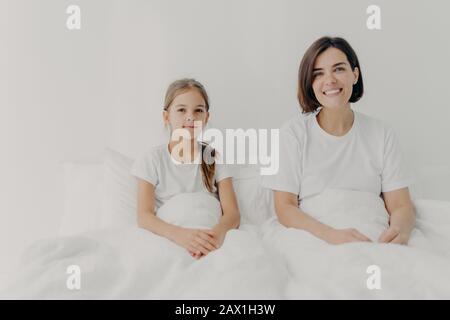 Horizontale Aufnahme von entspannter Mutter und Tochter im Bett unter weißer Decke, Blick in die Kamera, fühlen sich zufrieden, verbringen Sie freie Zeit am Morgen Stockfoto