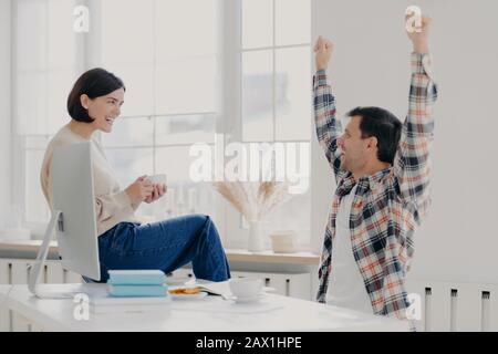 Glücklicher junger Mann freut sich über die erfolgreiche Arbeit, hebt die Arme, gekleidet in kariertem Hemd, froh sein, teilt seinen Erfolg mit Frau, die auf dem sitzt Stockfoto