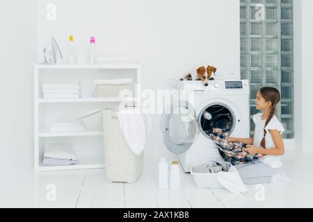 Haushaltskonzept, Kinder und Hausaufgaben. Das glückliche Kind entlädt die Waschmaschine, legt saubere, gewaschene Kleidung in das Becken, neugieriger Hund schaut aus dem Atem Stockfoto