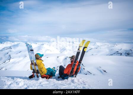 Frau und Mann sitzen gegen Skier mit Bergen im Hintergrund Stockfoto