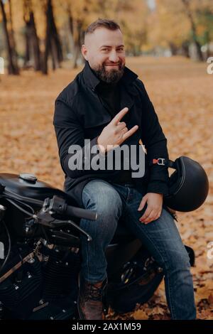 Der hübsche bärtige männliche Motorradfahrer hält den Helm, macht mit den Fingern Horn-Geste, fühlt sich cool an, trägt schwarzen Mantel und Jeans, sitzt wieder auf schnellem Motorrad Stockfoto