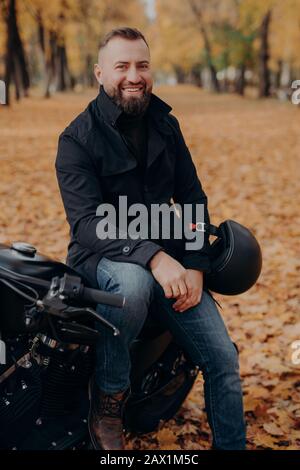 Vertikales Bild eines lächelnden Motorradfahrers lächelt fröhlich, hält Helm, posiert auf Motorrad, trägt schwarze Jacke und Jeans, ist durch Park gefahren, genießt Stockfoto