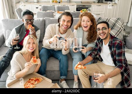 Eine Gruppe von fünf multiethnischen Freunden, die Spaß beim Essen von Pizza und beim Spielen von Videospielen haben, die auf die Kamera blicken, horizontale Aufnahme Stockfoto