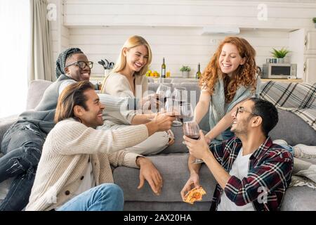 Horizontaler Schuss fröhlicher junger Männer und Frauen verbringen das Wochenende zusammen in einer gemütlichen Wohnung, in der Gläser mit Wein geklinkt sind Stockfoto