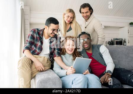 Die horizontale Aufnahme von fünf jungen Menschen versammelte sich im Wohnzimmer, um etwas Lustiges auf dem Tablet-PC zu sehen Stockfoto