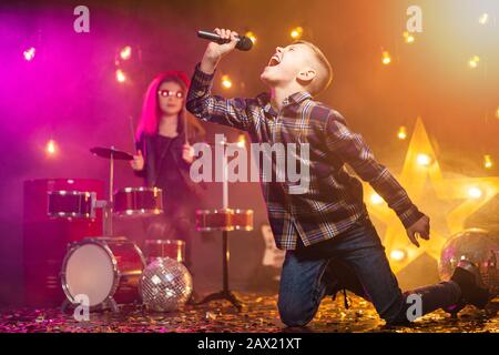 Kinder geben vor, eine Rockband zu sein und im Studio oder auf der Bühne zu singen. Junge singen und Mädchen zahlen am Schlagzeug Stockfoto