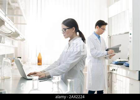 Horizontaler Schuss von jungen Männern und Frauen, die weiße Mäntel tragen, die im modernen Labor stehen und mit Dokumenten arbeiten Stockfoto