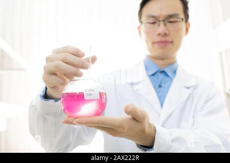 Horizontales Porträt des jungen Mediziners, der weißen Mantel und Brillen trägt, die den Kolben mit rosafarbener Flüssigkeit halten Stockfoto
