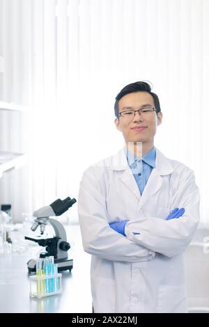 Selbstbewusster asiatischer Pharmakologe mit weißem Labormantel und Brillen, die mit gekreuzten Armen stehen und auf die Kamera blicken, die im modernen Labor lächelt Stockfoto