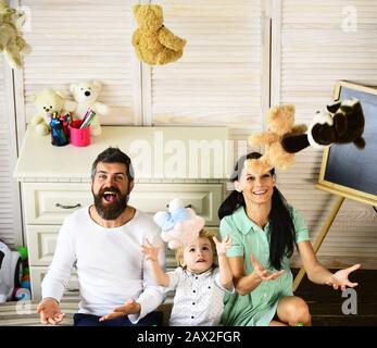 Mutter, Vater und Sohn sitzen auf dem Boden und werfen bunte Spielzeuge hoch. Eltern, die mit Teddybären spielen Stockfoto