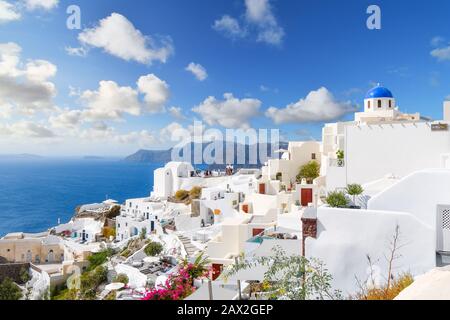 Oia, Santorini, Griechenland im Sommer. Weiß getünchte Häuser und blaue Kuppelkirche unterstreichen die Insel, während Touristen einen Blick auf die Terrasse des Resorts auf das Meer genießen. Stockfoto