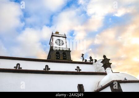Die Außenfassade der St. Sebastian Kirche, Igreja Matriz de Sao Sebastiao, in Ponta Delgada, Azoren, Portugal. Weißer Uhrturm von unten mit blauem Himmel und Wolken darüber. Wolken bei Sonnenuntergang.