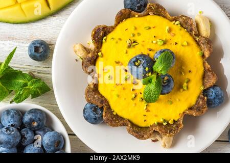 Veganes Dessert. Roher veganer gelber Mangokuchen mit frischen Blaubeeren, Pistazien und Minze. Gesunde, leckere glutenfreie Speisen. Draufsicht Stockfoto