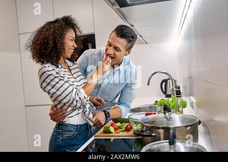 Romantisches Datum. Junges multiethnisches Paar, das in der Küche steht und seine Freundin zum Abendessen kocht, gibt dem Freund Gemüse zum lachen und verspielt Stockfoto